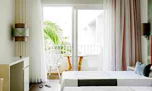 Vaalea hotellihuone, jossa on kaksi erillistä vuodetta ja parveke, jossa tuoleja ja pöytä. (Tekoälyn luoma teksti)