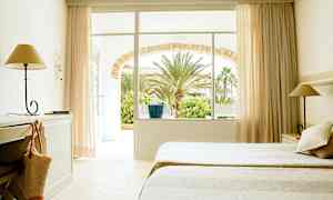 Hotellihuone, jossa on kaksi sänkyä, kirjoituspöytä ja lamppu. Ikkuna ja ovi johtavat puutarhaan, jossa on palmuja. (Tekoälyn luoma teksti)