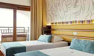 Hotellihuone, jossa on kaksi yhden hengen vuodetta turkooseilla koristeilla ja taustalla seinällä taiteellinen koristelu sekä pieni lamppu. (Tekoälyn luoma teksti)