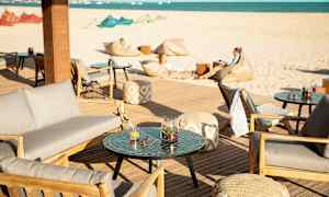 Ulkoterassi, jossa istumapaikkoja ja pöytiä, taustalla hiekkaranta ja meri. (Tekoälyn luoma teksti)