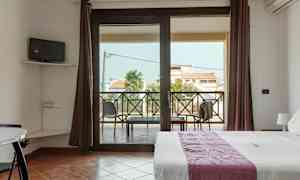 Huone, jossa on sänky, pieni televisio seinällä ja parveke, jolta on näkymä rakennuksiin ja palmuihin. (Tekoälyn luoma teksti)