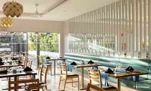 Moderni ja valoisa ravintolatila, jossa on pöytiä ja tuoleja, ja ikkunat avautuvat vehreään ulkoalueeseen. (Tekoälyn luoma teksti)