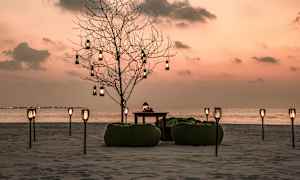 Rantaympäristö auringonlaskun aikaan, koristeltu puu, istuinpussit ja pöytä pitkien lyhtyjen joukossa. (Tekoälyn luoma teksti)