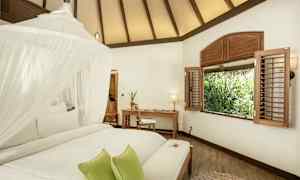 Vaalea huone, jossa on valkoinen katto, sänky, jossa hyttysverkko ja vihreät tyynyt, työpöytä ja tuoli ikkunan vieressä ja näkymä vehreyteen. (Tekoälyn luoma teksti)