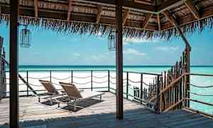 Kaksi aurinkotuolia puisella laiturilla, näkymä täynnä kirkasta sinistä merta olkikatoksen alla. (Tekoälyn luoma teksti)