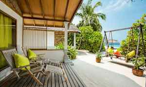 Ulkotila puutuoleilla, keinu ja hiekka, ympäröitynä vehreällä kasvillisuudella ja palmupuiden joukossa. (Tekoälyn luoma teksti)