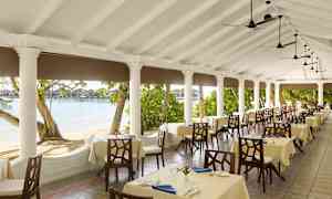 Ravintolan terassi, jossa on katettuja pöytiä, pylväiden ympäröimä ja näkymä rannalle ja vedelle. (Tekoälyn luoma teksti)