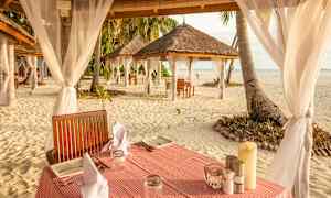 Pöydät katettu servieteillä ja lautasilla hiekkarannalla, ympäröitynä koristeellisilla kankailla varustetuilla katoilla. (Tekoälyn luoma teksti)