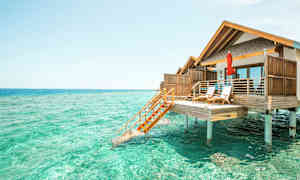 Valokuva bungalowista pylväiden päällä turkoosin veden yllä. (Tekoälyn luoma teksti)