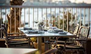 Pöytä on katettu ulkoterassilla, varusteltuna laseilla ja lautasliinoilla, ympärillään näkymä vedelle. (Tekoälyn luoma teksti)