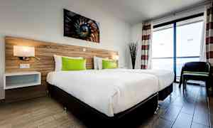 Hotellihuone, jossa on kaksi erillistä vuodetta, valkoiset vuodevaatteet ja vihreät tyynyt, puupaneelipääty sekä suuret ikkunat, jotka päästävät luonnonvaloa sisään. (Tekoälyn luoma teksti)