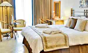 Hotellihuone, jossa on suuri sänky, vuodevaatteet ja tyynyt. Kaksi tuolia ja pöytä ovat sivulla, ja taustalla näkyy työskentelytila peileineen ja kirjoituspöytineen. (Tekoälyn luoma teksti)