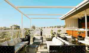 Kuvassa kattoterassi, jossa on istumaryhmiä, pöytiä ja varjoja. Näkymä rakennuksiin ja kirkkaansininen taivas. (Tekoälyn luoma teksti)