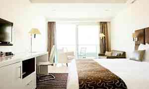 Hotellihuone, jossa on suuri sänky, kirjoituspöytä, tuoli, sohva ja parveke lasiovilla. (Tekoälyn luoma teksti)