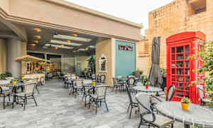 Ulkotila, jossa on pöytiä ja tuoleja kahvilassa. Punainen puhelinkioski ja vihreitä kasveja. (Tekoälyn luoma teksti)