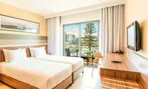 Hotellihuone, jossa kaksi yhden hengen vuodetta, työpöytä ja parvekenäkymä uima-altaalle ja puihin. (Tekoälyn luoma teksti)