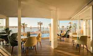 Ravintola, jossa on istumapaikkoja ja lasiseinät, joista on näkymä uima-altaalle ja palmuja meren äärellä. (Tekoälyn luoma teksti)