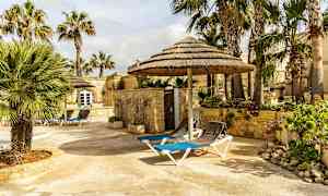 Kiveys, jossa on aurinkotuoleja ja oljesta katettuja varjoja, palmujen ja kivirakennusten ympäröimä. (Tekoälyn luoma teksti)