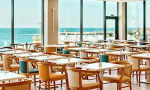 Ravintola, jossa on useita pöytiä ja tuoleja, suuri ikkunanäkymä merelle. (Tekoälyn luoma teksti)