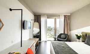 Moderni hotellihuone, jossa suuri sänky, istuinryhmä ikkunan vieressä ja näkymä satamaan sekä työpiste tuolilla. (Tekoälyn luoma teksti)