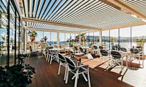 Ulkoilmaterassi, jossa on pöytiä ja tuoleja, lasiseinät ympärillä ja näkymä merelle sekä palmuihin. (Tekoälyn luoma teksti)