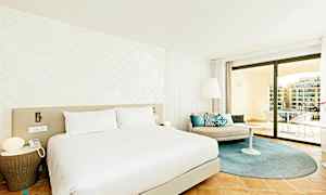 Moderni hotellihuone, jossa on suuri sänky, vaaleat seinät ja tyylikäs sisustus. Huoneessa on istuma-alue tyynyineen sekä tilava parveke näkymillä. (Tekoälyn luoma teksti)