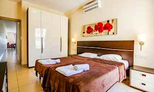 Makuuhuone, jossa on kaksi erillistä vuodetta, ruskeat päiväpeitot ja valkoiset vuodevaatteet. Seinällä sänkyjen yläpuolella roikkuu taulu, jossa on punaisia kukkia. Vasemmalla on valkoinen kiiltävä vaatekaappi. (Tekoälyn luoma teksti)