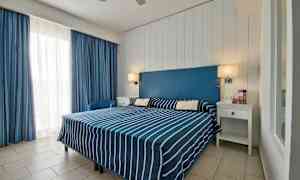 Hotellihuone, jossa on parisänky sinivalkoraidallisilla vuodevaatteilla, sininen sängynpääty ja valkoiset huonekalut. (Tekoälyn luoma teksti)