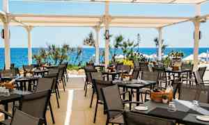 Terassi ulkoravintolan istumapaikoilla ja pöydillä valkoisen katon alla, meri ja kasvillisuus taustalla. (Tekoälyn luoma teksti)