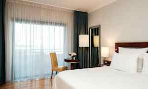 Hotellihuone, jossa on iso sänky, pöytä ja tuoli sekä ikkuna valkoisilla verhoilla. (Tekoälyn luoma teksti)