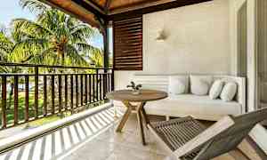 Terassi, jossa on valkoinen sohva, pyöreä puupöytä ja aurinkotuoli, näkymä palmupuihin ja vihreä alue. (Tekoälyn luoma teksti)