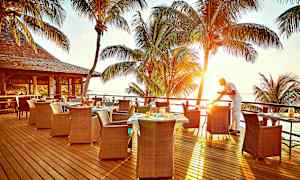 Ulkoterassilla näkymä merelle ja auringonlaskuun, taustalla palmupuita ja tarjoilija valmistelee pöytää. (Tekoälyn luoma teksti)