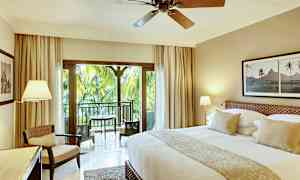 Tyylikkäästi sisustettu hotellihuone, jossa iso sänky, istuinalue ja parveke, jolta on näkymä vehreisiin palmuihin. (Tekoälyn luoma teksti)