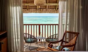 Näkymä merelle huoneesta, jossa on katettu parveke. Läpinäkyvien verhojen välissä on kaksi puutarhatuolia pöydän ääressä. (Tekoälyn luoma teksti)