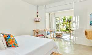 Vaalea hotellihuone, jossa on sänky, kirjoituspöytä ja istuinryhmä. Suuret ikkunat, joista avautuu näkymä vehreään puutarhaan. (Tekoälyn luoma teksti)