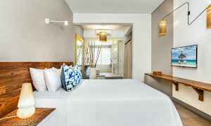 Kuva modernista hotellihuoneesta, jossa on puukalusteet, sänky valkoisilla lakanoilla ja värikkäillä tyynyillä sekä suuri ikkuna taustalla. (Tekoälyn luoma teksti)