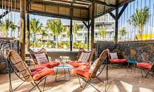 Puutarhassa sijaitseva ulkotila, jossa korituoleja ja värikkäitä tyynyjä sekä pöytä hiekkapohjalla. Taustalla näkyy puu- ja kasvillisuusmaisemia. (Tekoälyn luoma teksti)