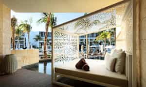 Ulkotiloihin sijoitettu lepotuoli, jossa on valkoiset tyynyt ja ympärillä valkoinen koristeellinen rakenne. Taustalla näkyy palmuja, uima-allas ja moderneja rakennuksia. (Tekoälyn luoma teksti)