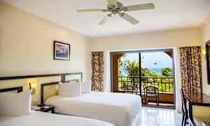 Hotellihuone, jossa kaksi sänkyä, kattotuuletin ja parveke, josta on näkymä trooppisiin palmuihin ja merelle. (Tekoälyn luoma teksti)