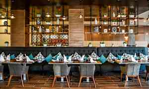 Tyylikäs ravintola, jossa on moderni sisustus. Pöydät on katettu laseilla ja lautasliinoilla, ja sohvat on koristeltu tyynyillä. Taustalla näkyy hyllyjä, joilla on koriste-esineitä ja kasveja. (Tekoälyn luoma teksti)