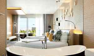 Moderni hotellihuone, jossa on suuri parivuode, koristeelliset tyynyt ja seinäkoristeita. Tilassa on erillinen kylpyamme sekä huonekaluja, kuten pöytiä ja tuoleja, lähellä lasiovea, josta on näkymä. (Tekoälyn luoma teksti)