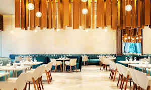 Moderni ravintola, jossa on tyylikkäitä tuoleja ja pöytiä. Koristeelliset valaisimet riippuvat katosta. (Tekoälyn luoma teksti)