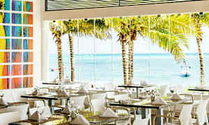 Ravintolatila, jossa on katettuja pöytiä ja suuret lasi-ikkunat, joista näkyy palmuja ja meri ulkopuolella. (Tekoälyn luoma teksti)