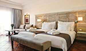 Hotellihuone, jossa on kaksi erillistä sänkyä, sohva, pöytä kukkamaljakolla ja ikkunalla verhot. (Tekoälyn luoma teksti)