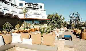 Ulkona sijaitseva oleskelualue, jossa on istuinryhmiä, tyynyjä ja kasveja, taustalla nykyaikainen rakennus. (Tekoälyn luoma teksti)