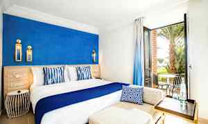 Vaalea hotellihuone, jossa on sininen korostusseinämä, iso sänky valkoisella ja sinisellä sisustuksella sekä avoin ovi, josta näkyy terassi tuoleineen ja palmuineen. (Tekoälyn luoma teksti)