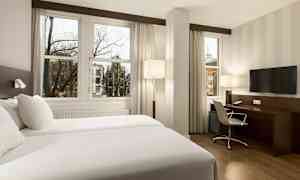 Hotellihuone, jossa on kaksi erillistä vuodetta, kirjoituspöytä, tuoli ja televisio, sekä isot ikkunat kaupunkimaisemaan. (Tekoälyn luoma teksti)