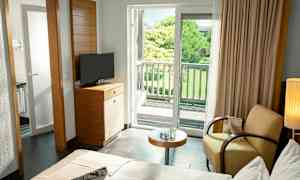 Hotellihuone, jossa on sänky, nojatuoli, pöytä, televisio ja parveke, jolta avautuu näkymä vehreään puutarhaan. (Tekoälyn luoma teksti)