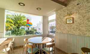 Kahvila, jossa pyöreä pöytä ja tuoleja ikkunan lähellä; ikkunasta näkyy palmupuu ja rakennus. (Tekoälyn luoma teksti)