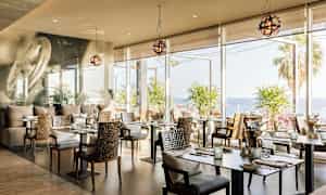 Ravintola, jossa on modernit pöydät ja tuolit, suuret ikkunat merelle ja palmuja. (Tekoälyn luoma teksti)